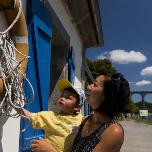 Jeune femme avec son bébé à près d'une maison éclusière arborant une bouée de sauvetage au bord d'un canal avec randonneurs au loin (voir l'image en plus grand)